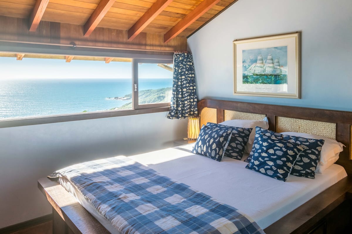 A bedroom with a bed, pillows, a framed picture, and a view of the ocean.
