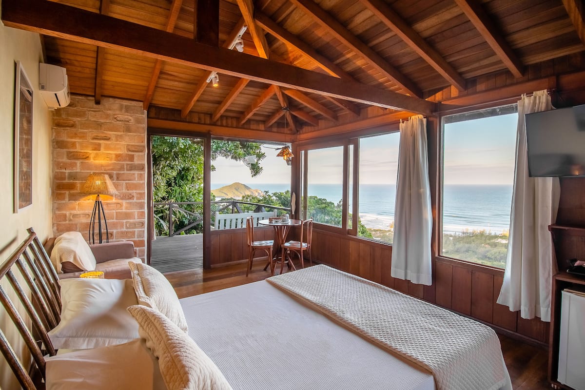 A bedroom with a large bed, wooden ceiling beams, a brick wall, a window with a view of the ocean, and a TV on a wooden stand.