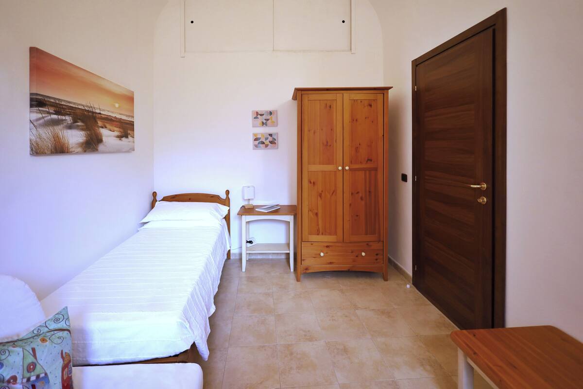 A bedroom with two single beds, a wooden wardrobe, a desk with a chair, and a large door.