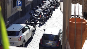 A narrow street lined with parked cars and motorcycles, with buildings featuring classical architecture and a prominent church in the background. - Cataneasy Rooms (Catania)