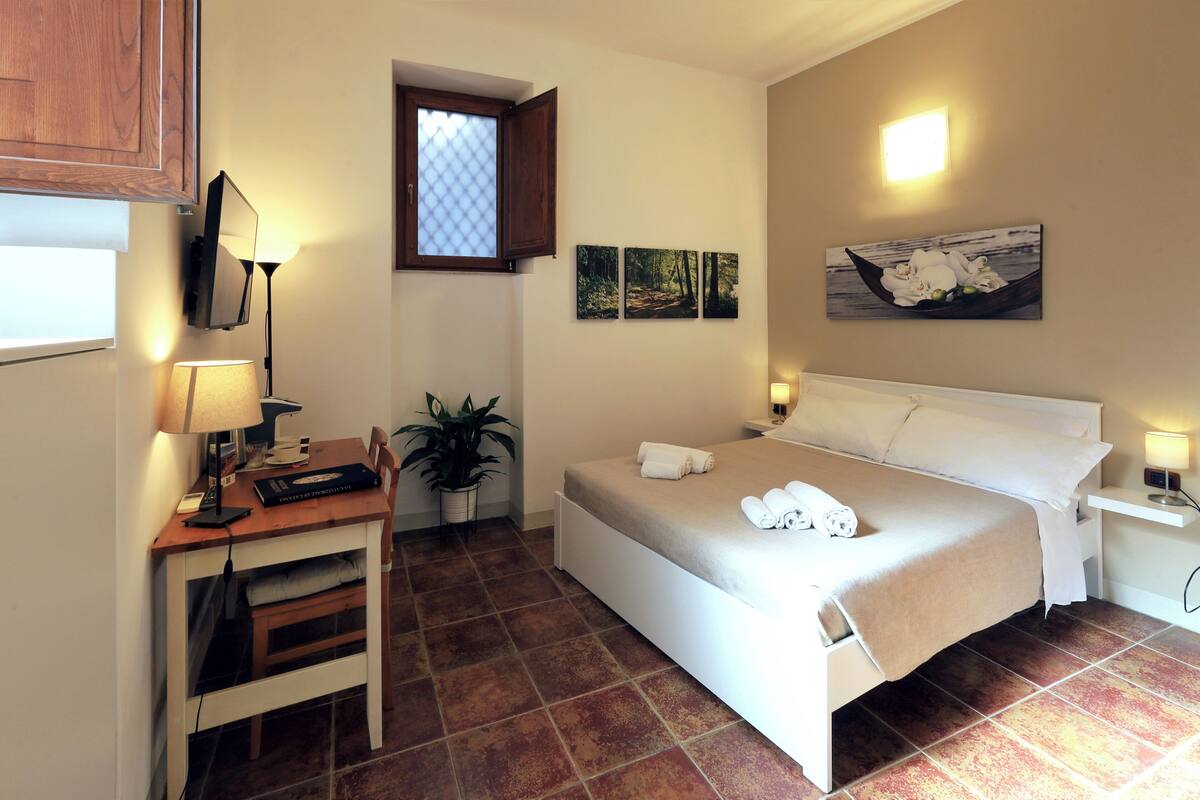 A bedroom with a bed, a desk with a computer, a chair, a potted plant, and a window with an open shutter.