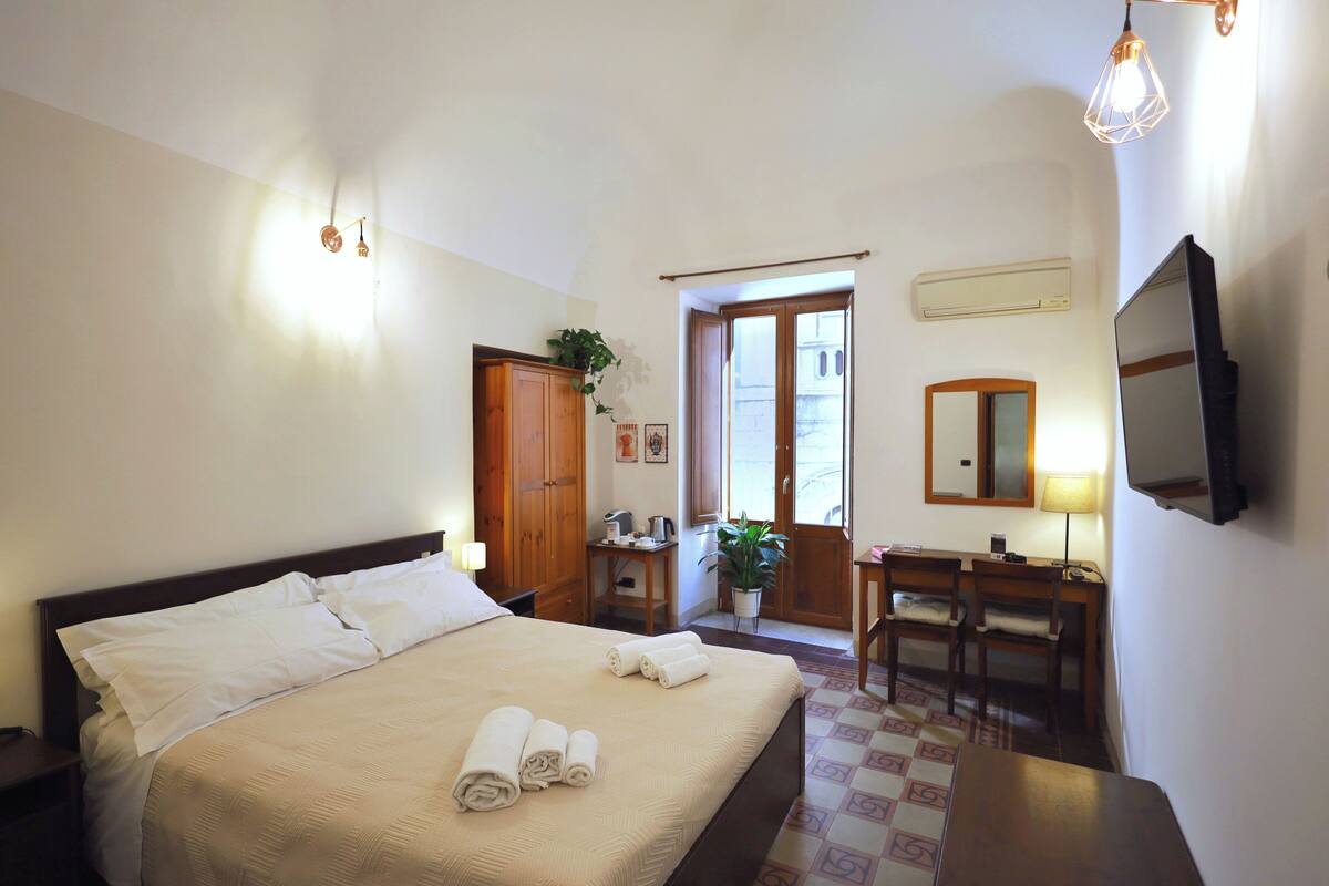 A hotel room with a bed, a desk, a chair, a TV, and a door leading to a balcony.