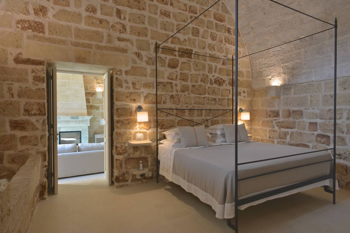 Un dormitorio con pared de piedra, una cama con dosel, lámparas de noche y un acceso a otra habitación.