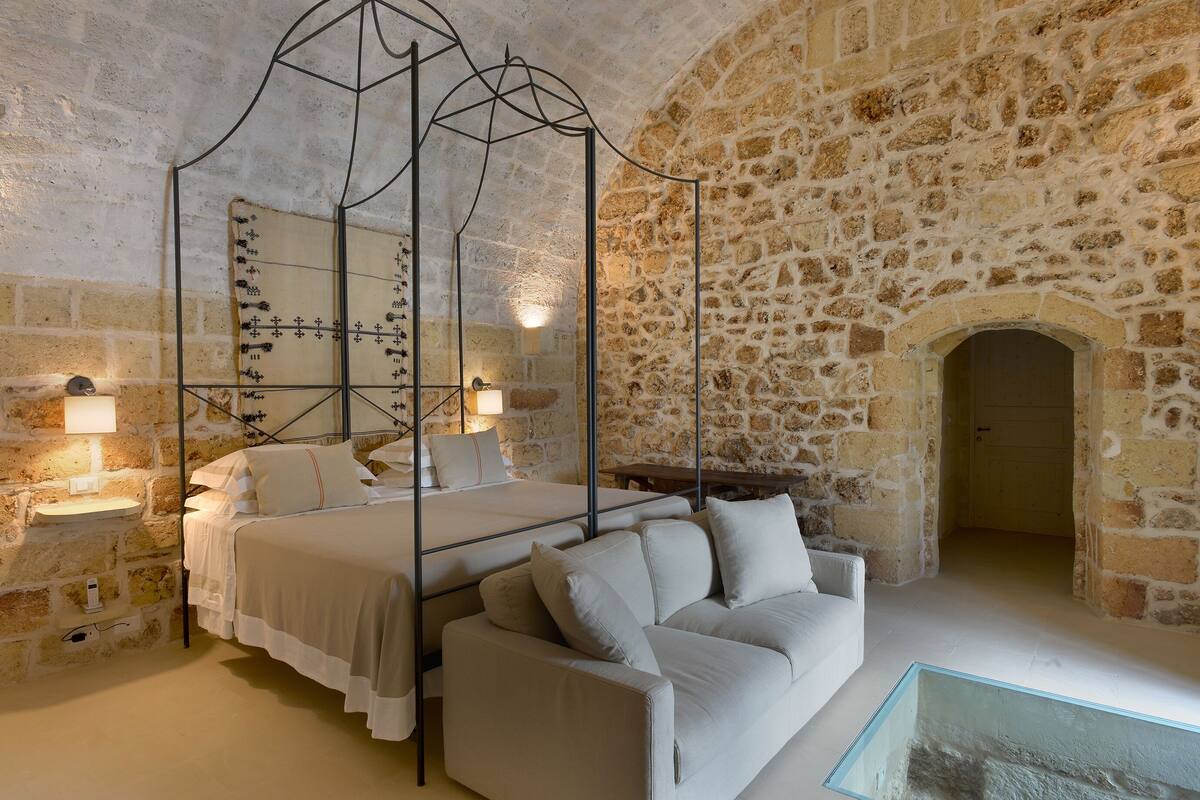 Un dormitorio con pared de piedra, cama con dosel, sofá y una mesa de centro de vidrio.