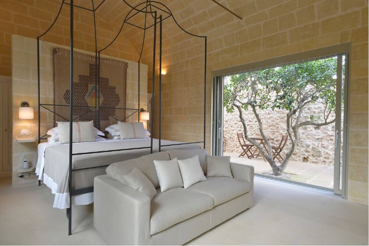 Un dormitorio con cama con dosel, un sofá y un amplio ventanal con vista a un árbol y zona de estar exterior.