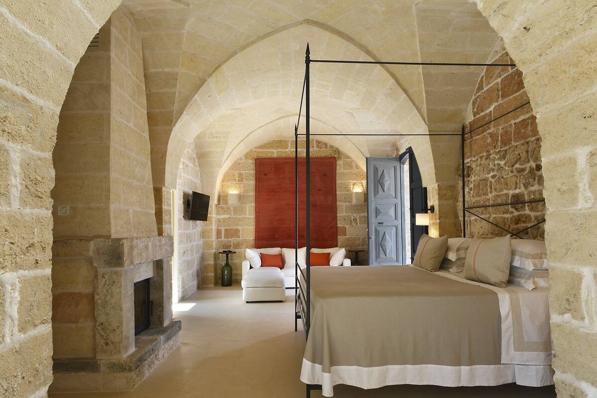Un dormitorio con cama con dosel, paredes de piedra, chimenea y una puerta roja.