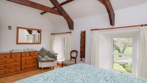 Une chambre à coucher avec un lit, une commode en bois, une chaise et une grande fenêtre donnant sur un paysage verdoyant.