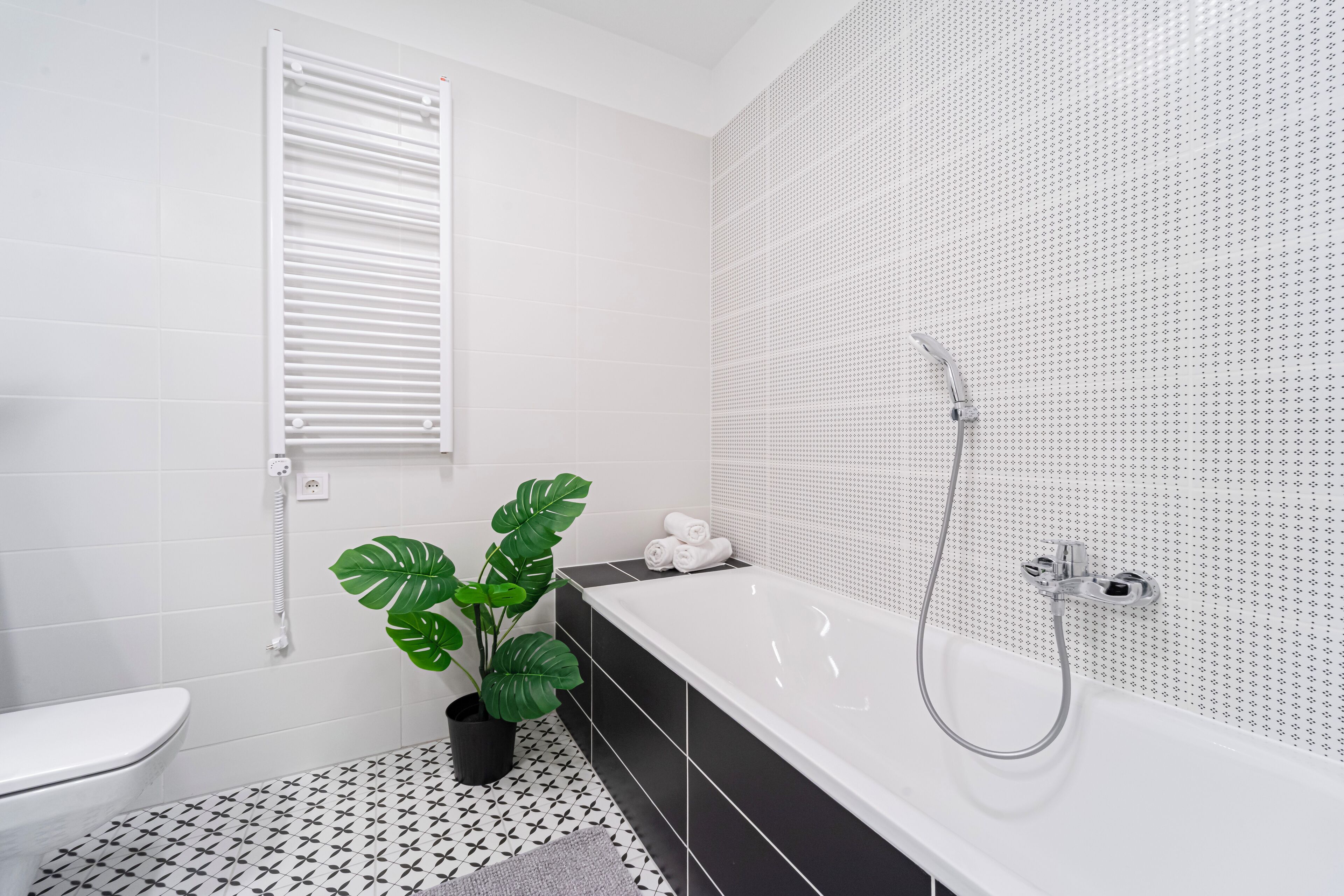 A modern bathroom with a white bathtub, black and white patterned floor tiles, a potted plant, and a white toilet.