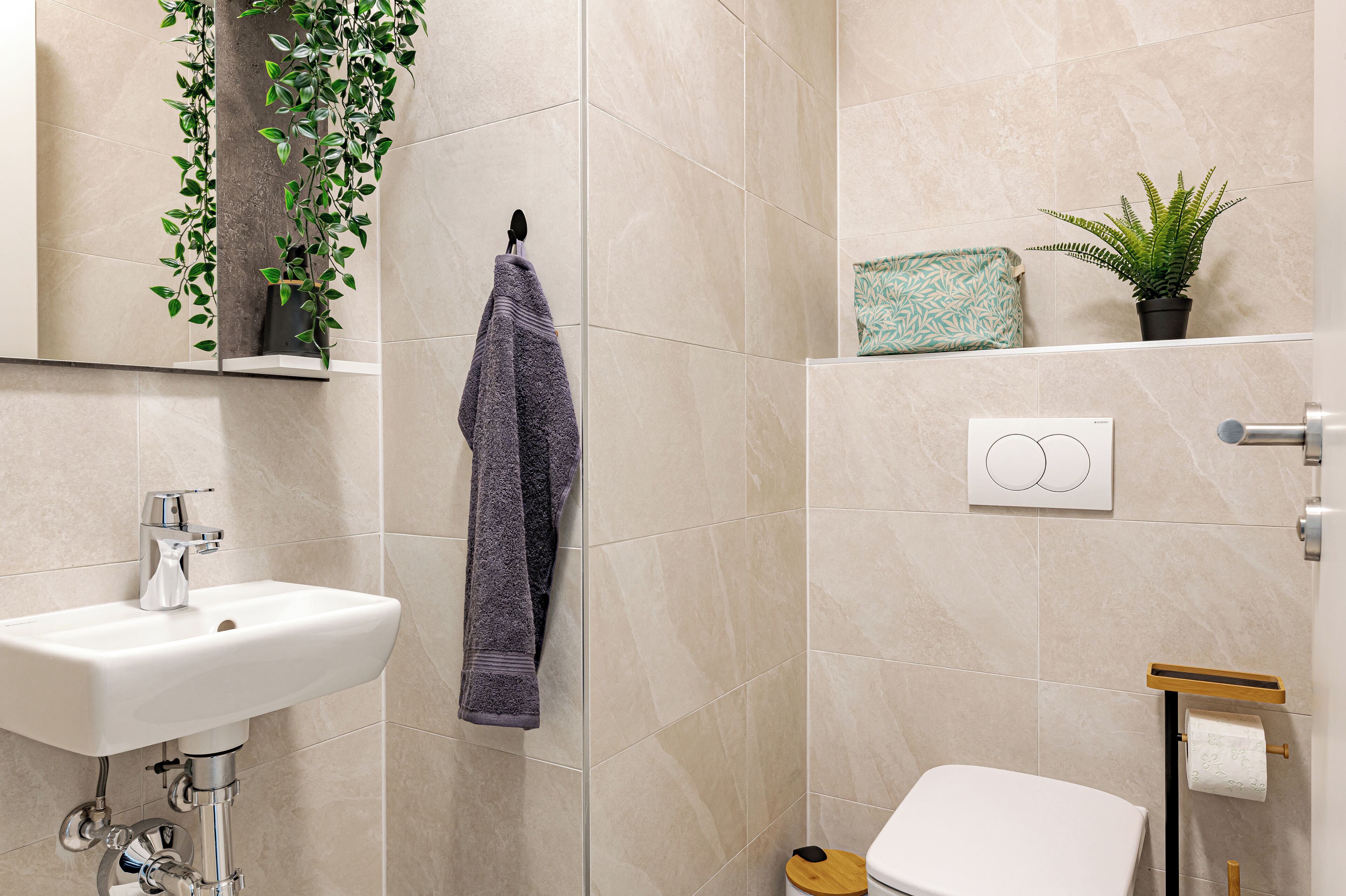 A bathroom with a white sink, a towel hanging on the wall, a toilet, and a small plant on the shelf.