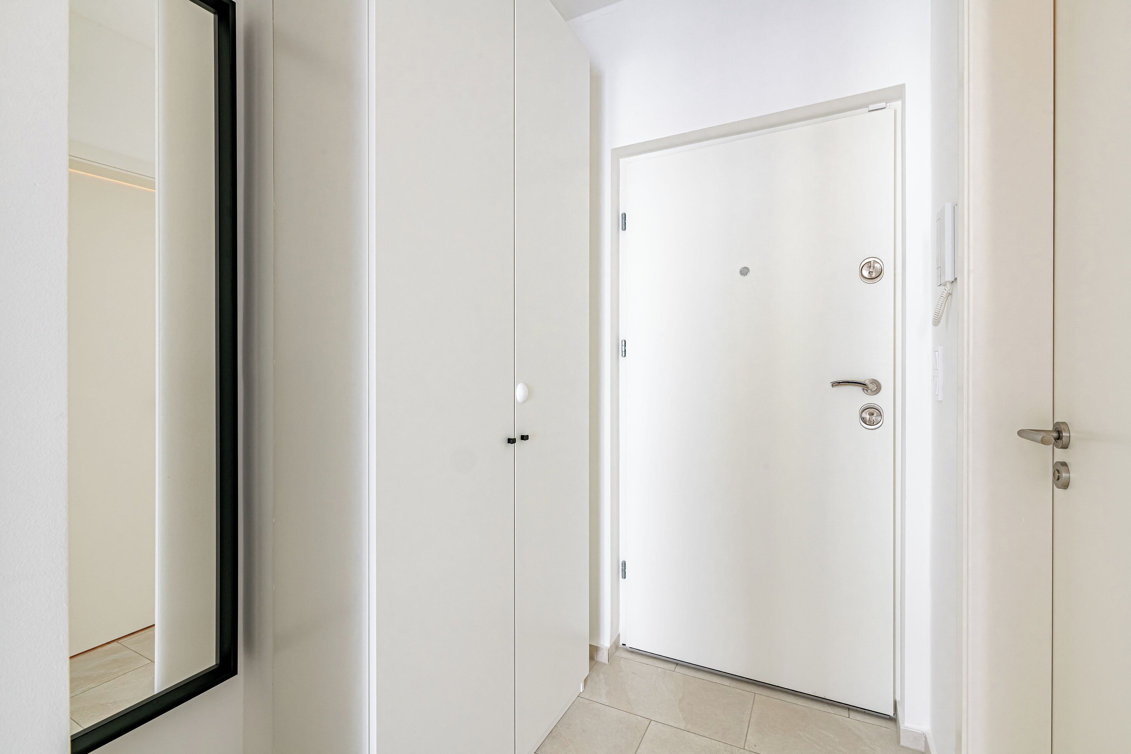 Une porte blanche avec un trou de serrure et une serrure, un miroir qui reflète une partie de la pièce et un sol carrelé.