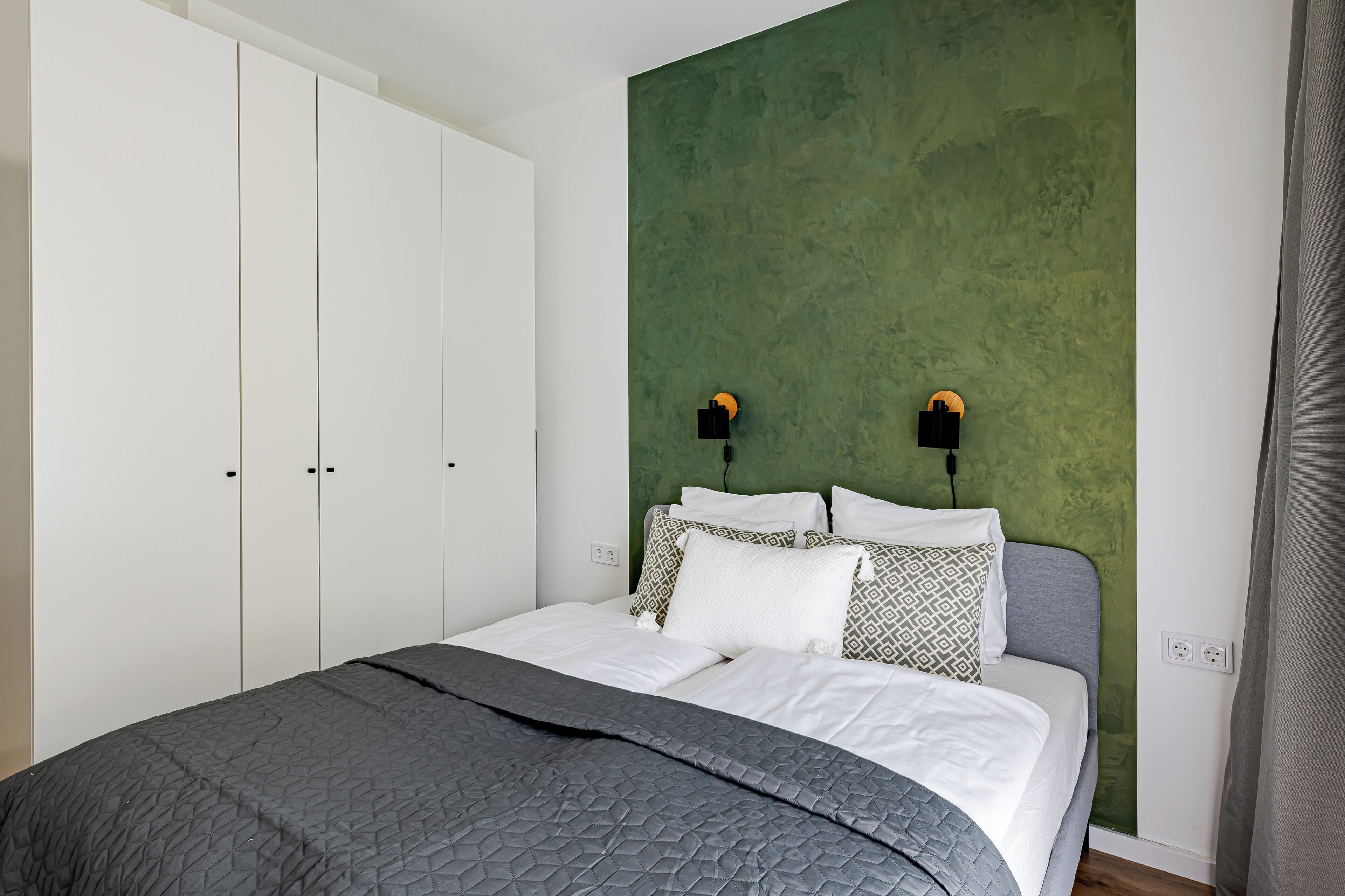A bedroom with a bed, white wardrobe, green accent wall, two wall-mounted lamps, and a grey curtain.