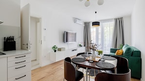 Una cocina moderna con armarios blancos, una zona de comedor con una mesa redonda y sillas, un sofá verde y un televisor montado en la pared.