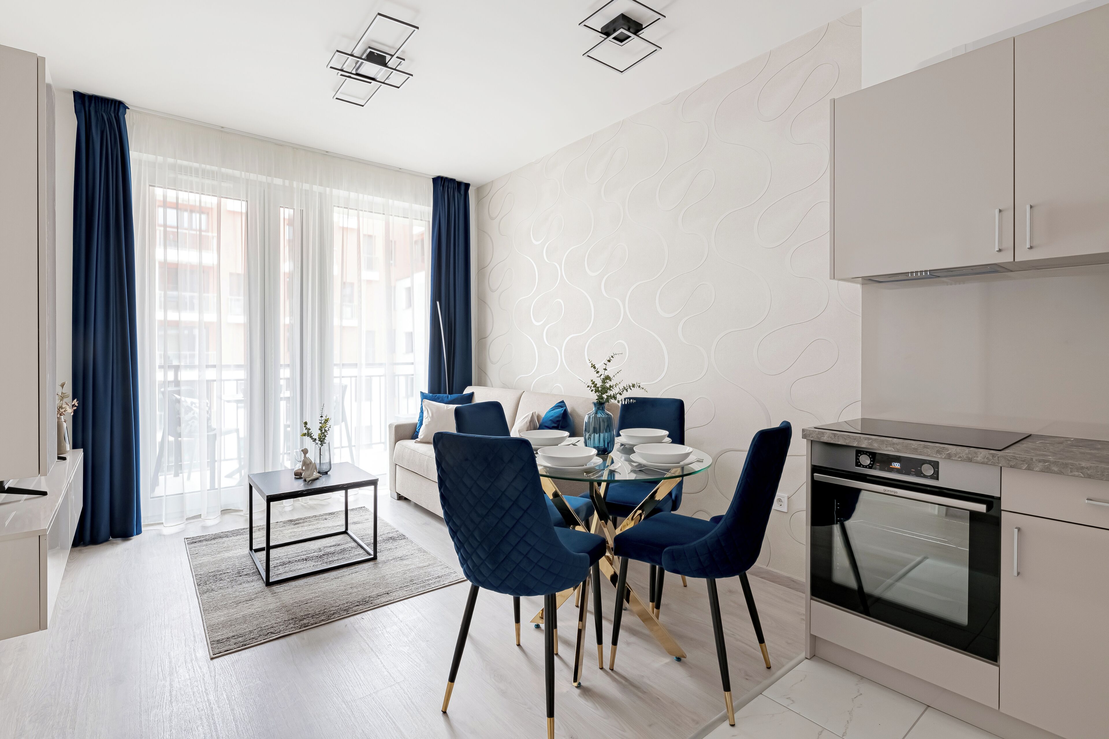 Cozinha moderna com forno embutido, área de refeições com mesa redonda e cadeiras azuis, e sala de estar com sofá e cortinas.