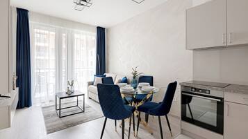 Cozinha moderna com forno embutido, área de refeições com mesa redonda e cadeiras azuis, e sala de estar com sofá e cortinas.
