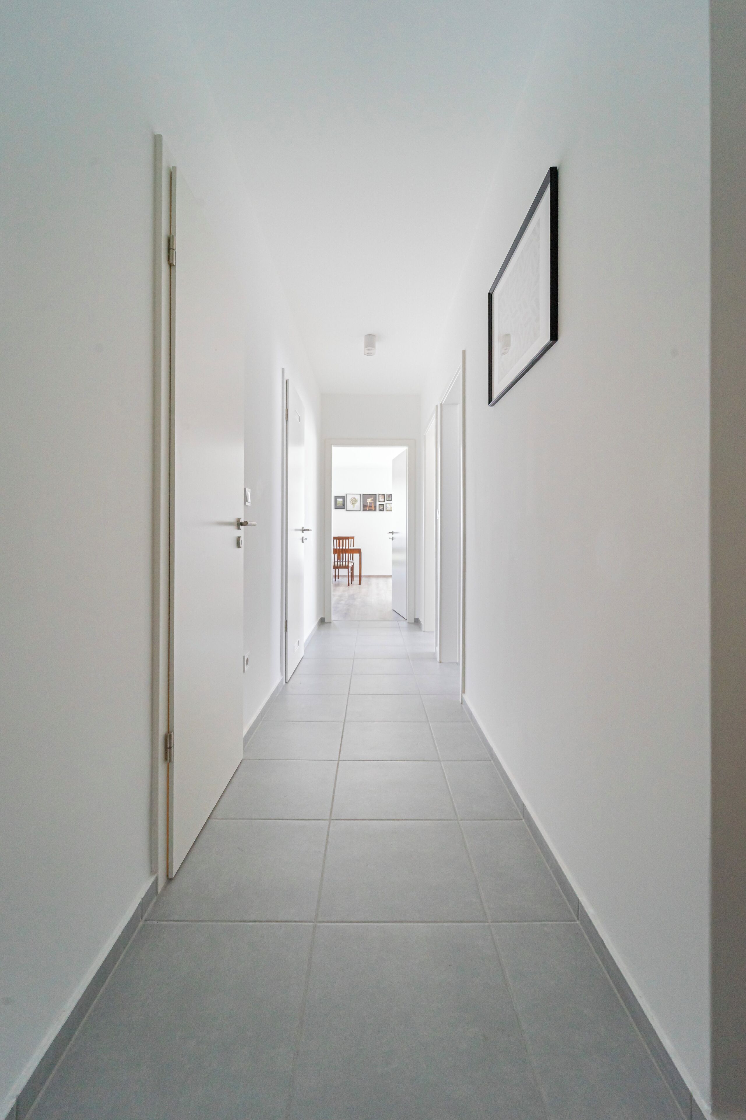 A long, narrow hallway with white walls and a tiled floor, leading to a room with a door at the end.
