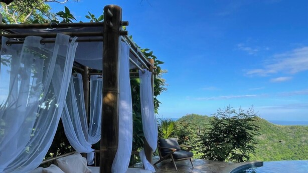 Un espace extérieur privé avec une terrasse en pierre, un coin salon couvert agrémenté de rideaux blancs, et une vue sur un paysage verdoyant et luxuriant.