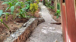 Un sentier en bois, abrité, qui traverse un jardin luxuriant, bordé de pierres et doté d’un feuillage dense.