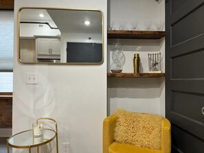 A modern interior with a mirror, a small round table with a candle, a yellow armchair, and a shelf with decorative items.