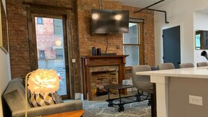 A modern living room with a fireplace, a television mounted on the wall, and a dining area with a cart. - Timeless Loft -OTR -TQL w/ FREE Parking (Cincinnati)