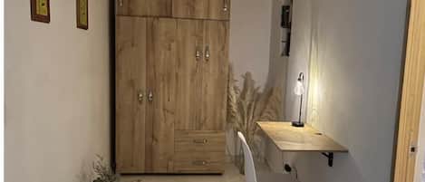Una habitación pequeña con un armario de madera, un escritorio con una silla, una lámpara y una planta pequeña.
