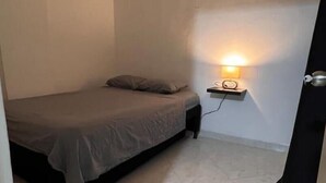 A small, sparsely furnished room with a bed, a bedside table with a lamp, and a tiled floor.