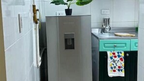 A compact kitchen with white tiles, a stainless steel refrigerator, a teal-colored sink, and a small potted plant on the fridge.
