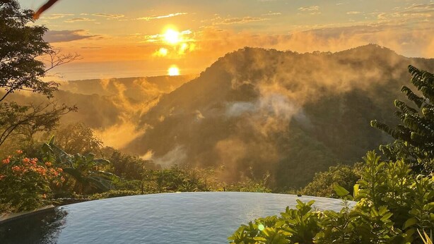 Une piscine à débordement offrant une vue sur un coucher de soleil au-dessus de montagnes brumeuses.