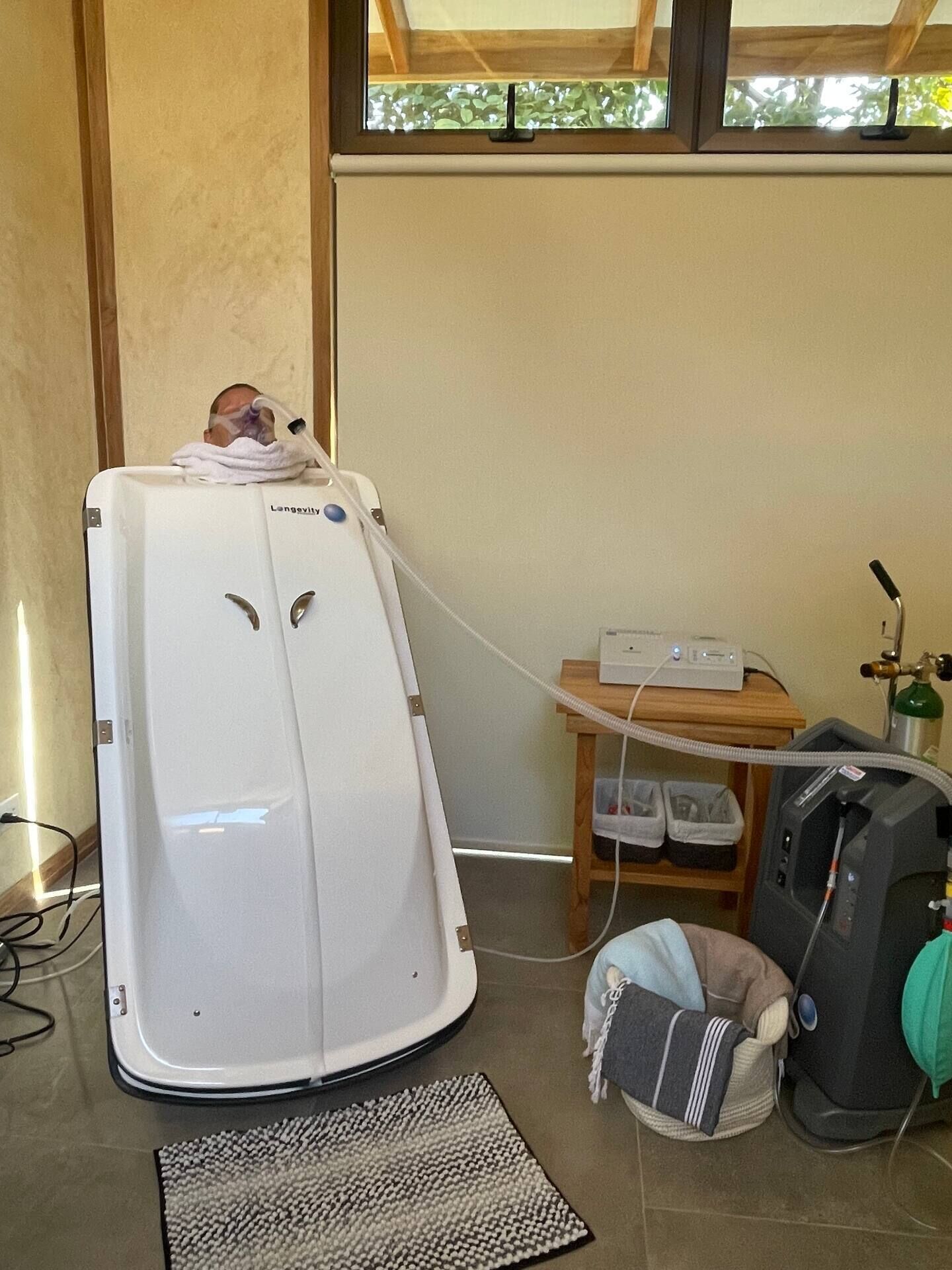 Un equipo médico, posiblemente una cámara hiperbárica, en una habitación con ventana, una mesa de madera y una bolsa en el suelo.