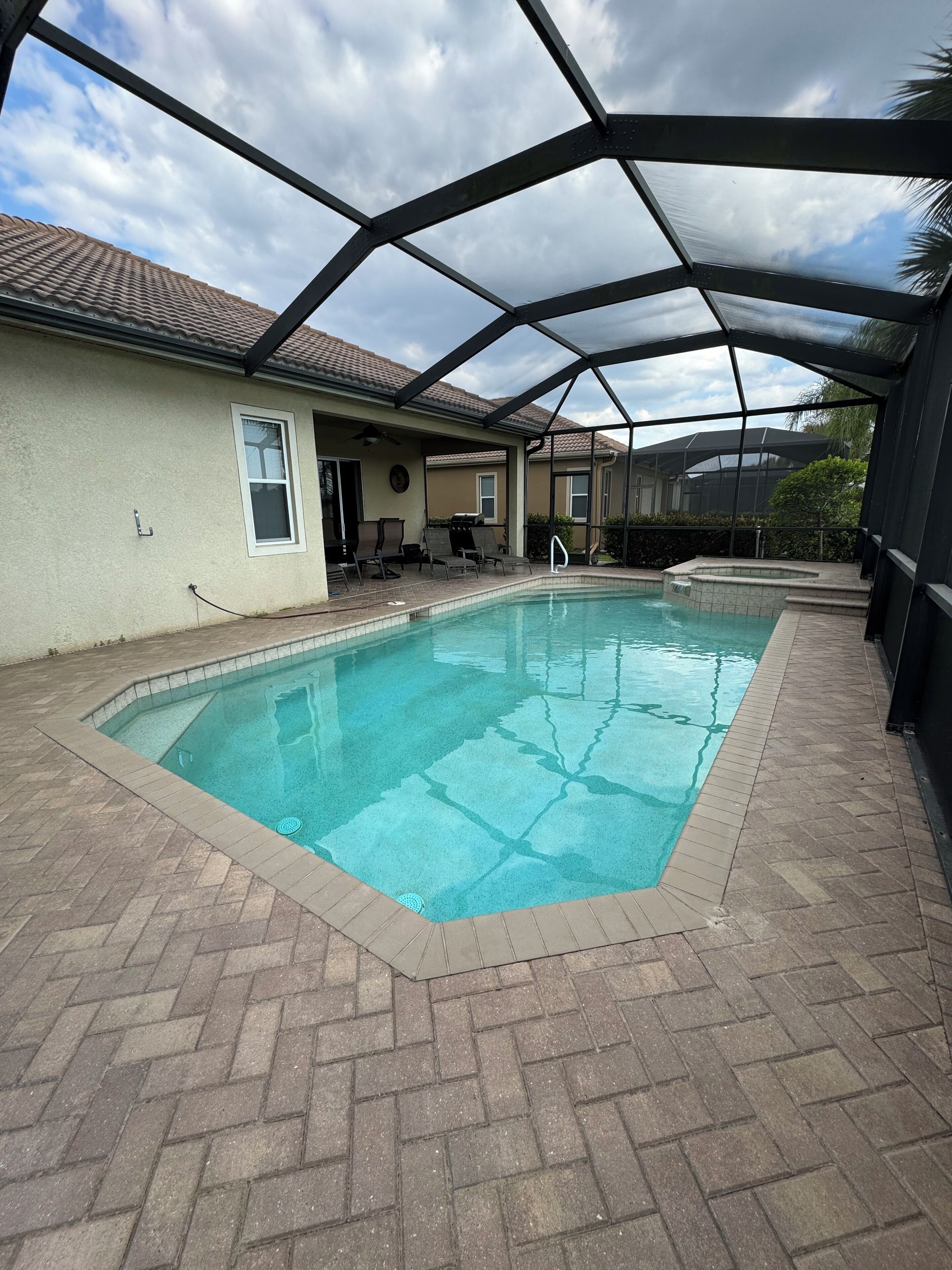 A covered outdoor swimming pool with a tiled deck, a covered patio area with chairs, and a view of neighboring houses.