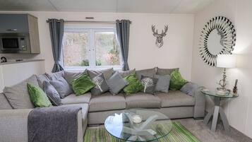 A modern living room with a grey sofa, green and grey cushions, a glass coffee table, a round side table with a lamp, and a decorative deer head on the wall.