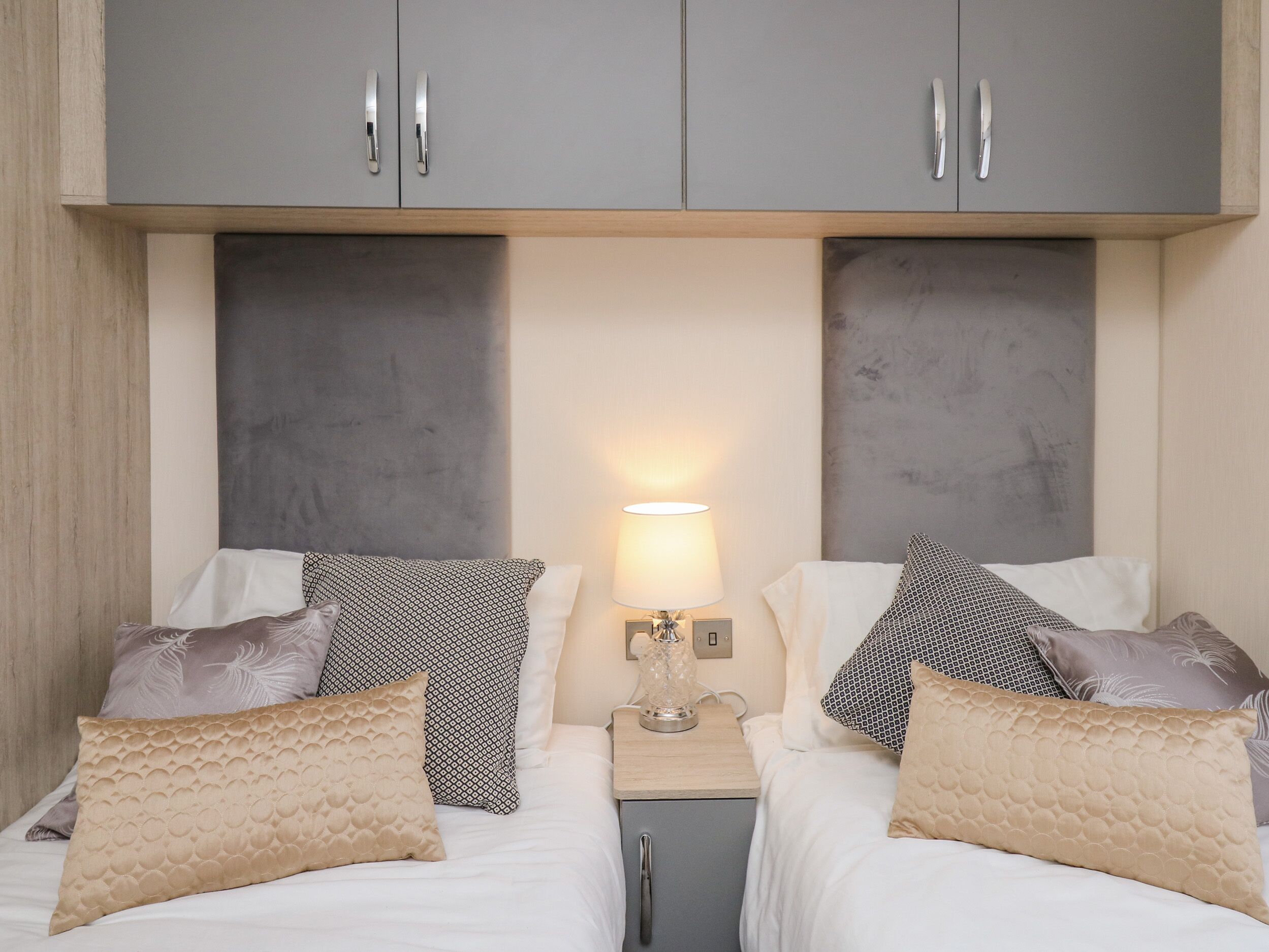 A hotel room with two beds, grey cabinets above, a bedside table with a lamp, and patterned pillows.