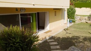 A modern building with a yellow facade, large sliding glass doors, a small garden area with a lawn and a rock border, and a visible outdoor area with a fence.