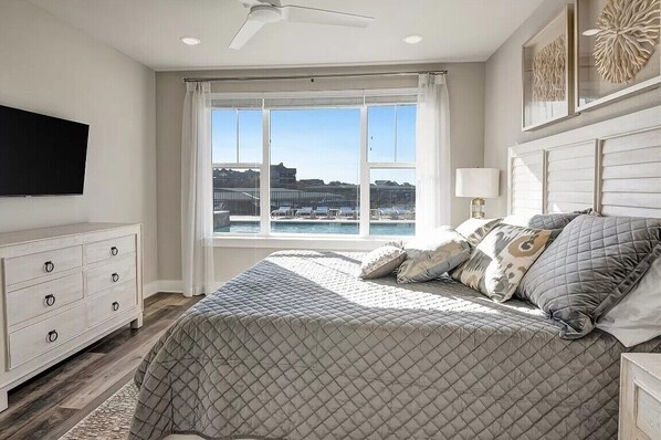 A bedroom with a large bed, a ceiling fan, a TV mounted on the wall, and a window with a view of a building and water. - NEW! First Floor Pool side PK Lake Luxury Condo - Hot Tub, FirePit, Gym, Laundry (Graford)