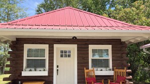 A log cabin with a red metal roof, two windows with white frames, a white door, and two rocking chairs with red cushions.