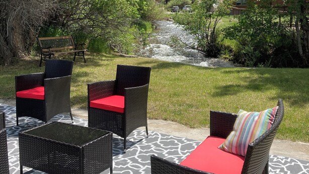 An outdoor seating area with wicker furniture and red cushions, surrounded by lush greenery and a view of a house and a flowing stream.