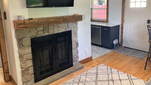 A living room with a stone fireplace, a mounted TV, and a kitchen area with a dishwasher.