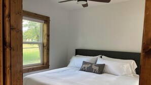 A bedroom with a bed, white bedding, a grey pillow with a moose design, a window with a view of greenery, and a ceiling fan.