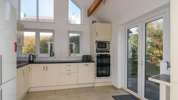 A modern kitchen with white cabinets, a black countertop, a microwave, and an oven. There are windows providing natural light and a view of the outside. - Tegfan (Newport)