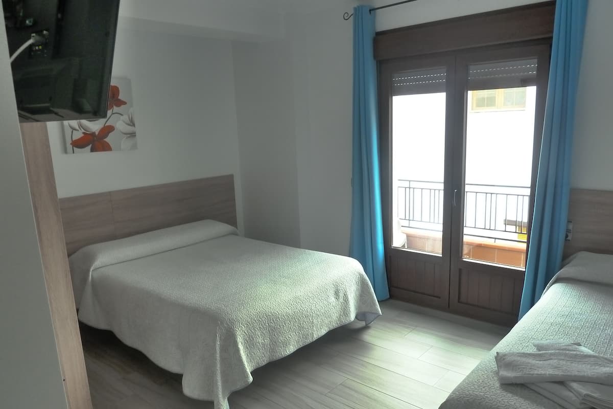 Ein Hotelzimmer mit zwei Einzelbetten, Holzboden, einem Balkon mit blauen Vorhängen und Blick nach draußen.