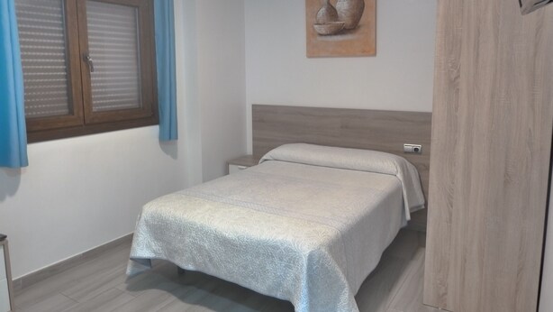 A bedroom with a bed, a nightstand, a wooden wardrobe, a window with curtains, and a painting on the wall.