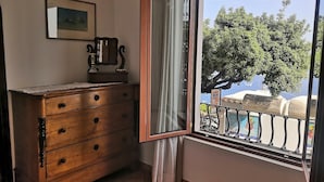 A room with a wooden dresser, a window with a view of a balcony and trees, and a framed picture on the wall. - Sea View in Positano 1 (Positano)