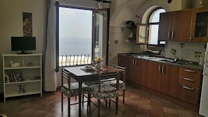 A kitchen with a dining table and chairs, a TV on a shelf, and a view of the sea through a window. - Sea View in Positano 1 (Positano)