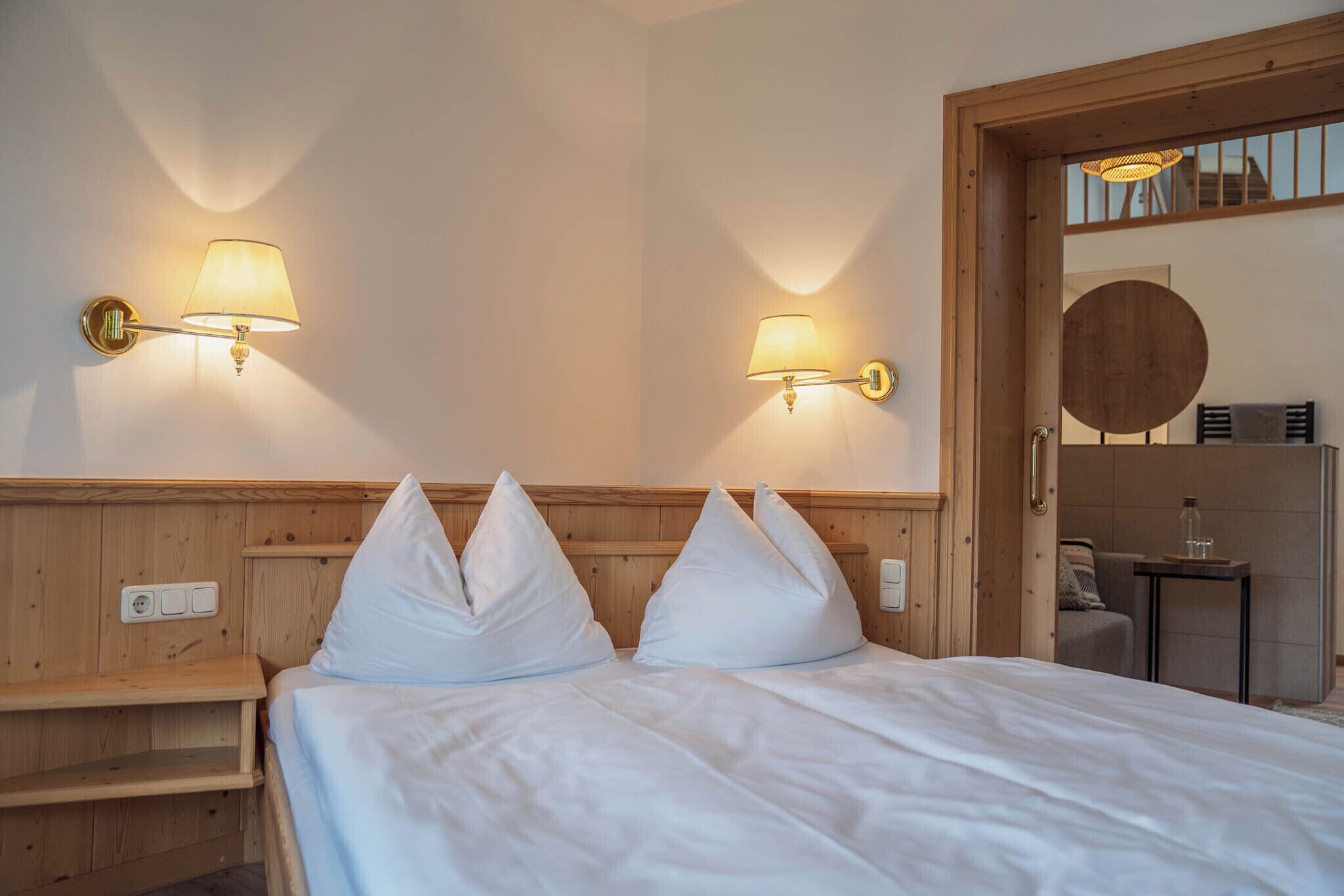 A hotel room with a wooden headboard, two bedside lamps, a bed with white bedding, and a small table with a glass.