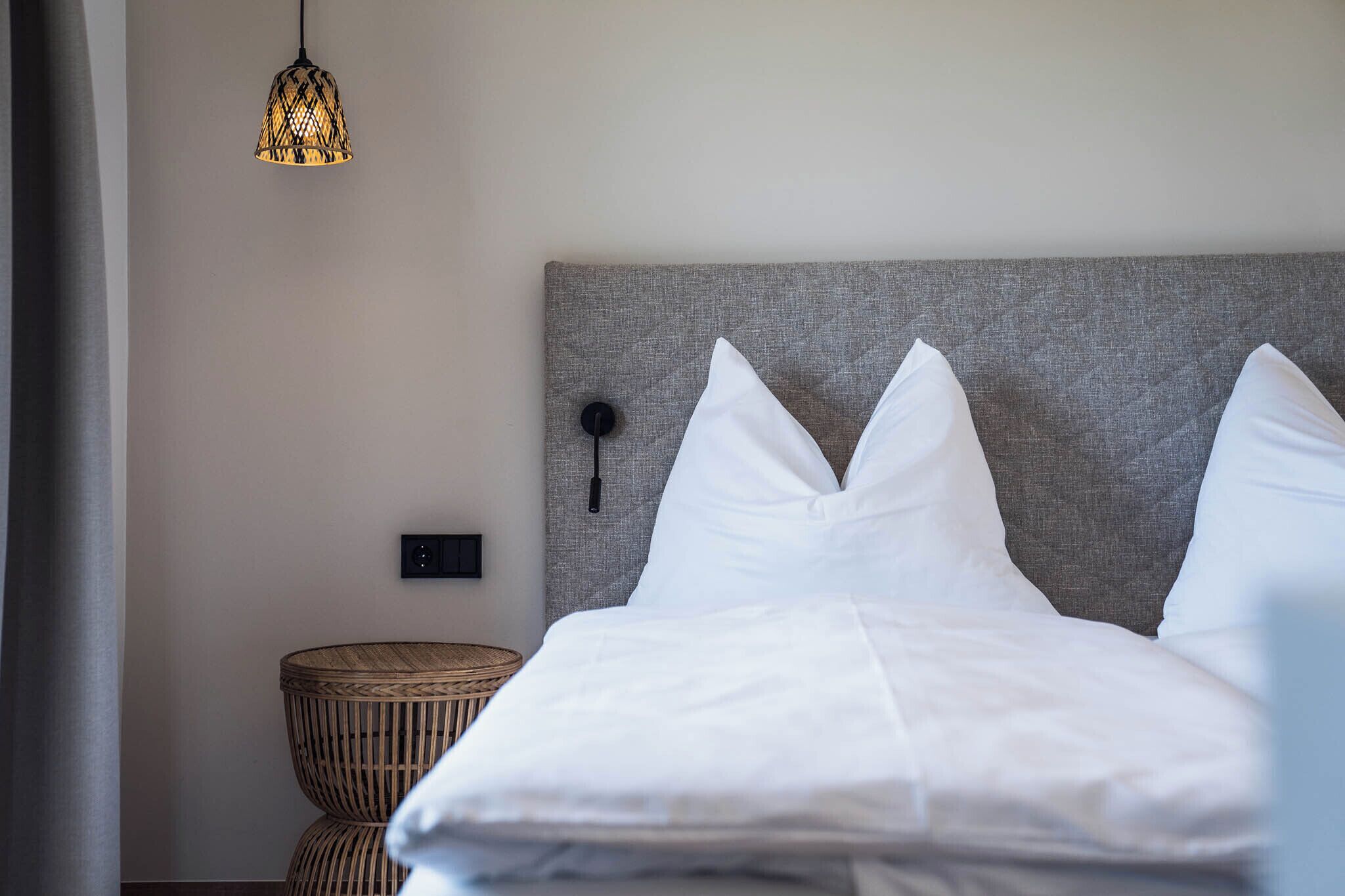 A neatly made bed with white linens, a grey headboard, and a woven side table.