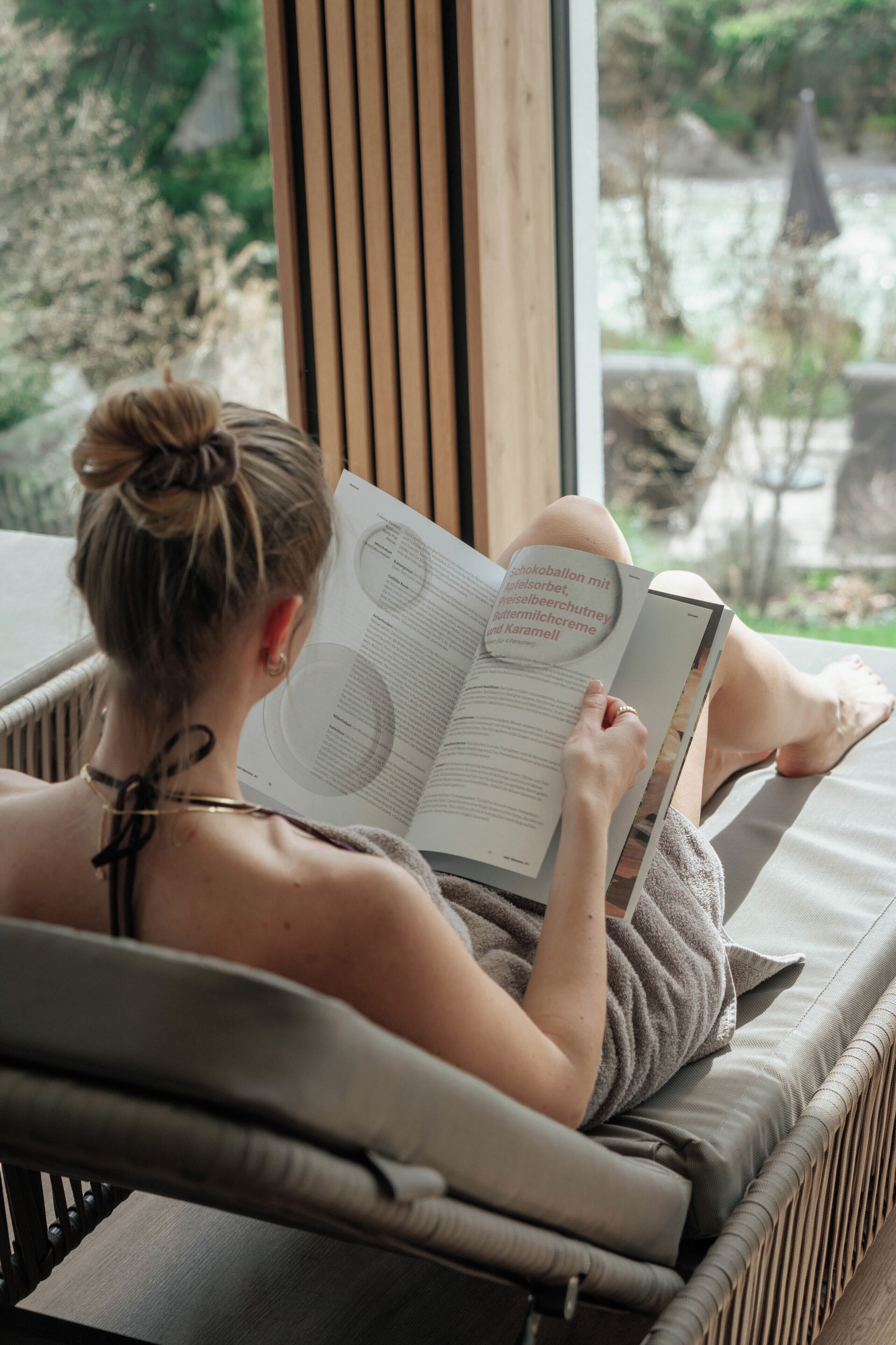 Una persona sentada en una silla de mimbre leyendo un libro junto a una ventana con vistas al verdor exterior.