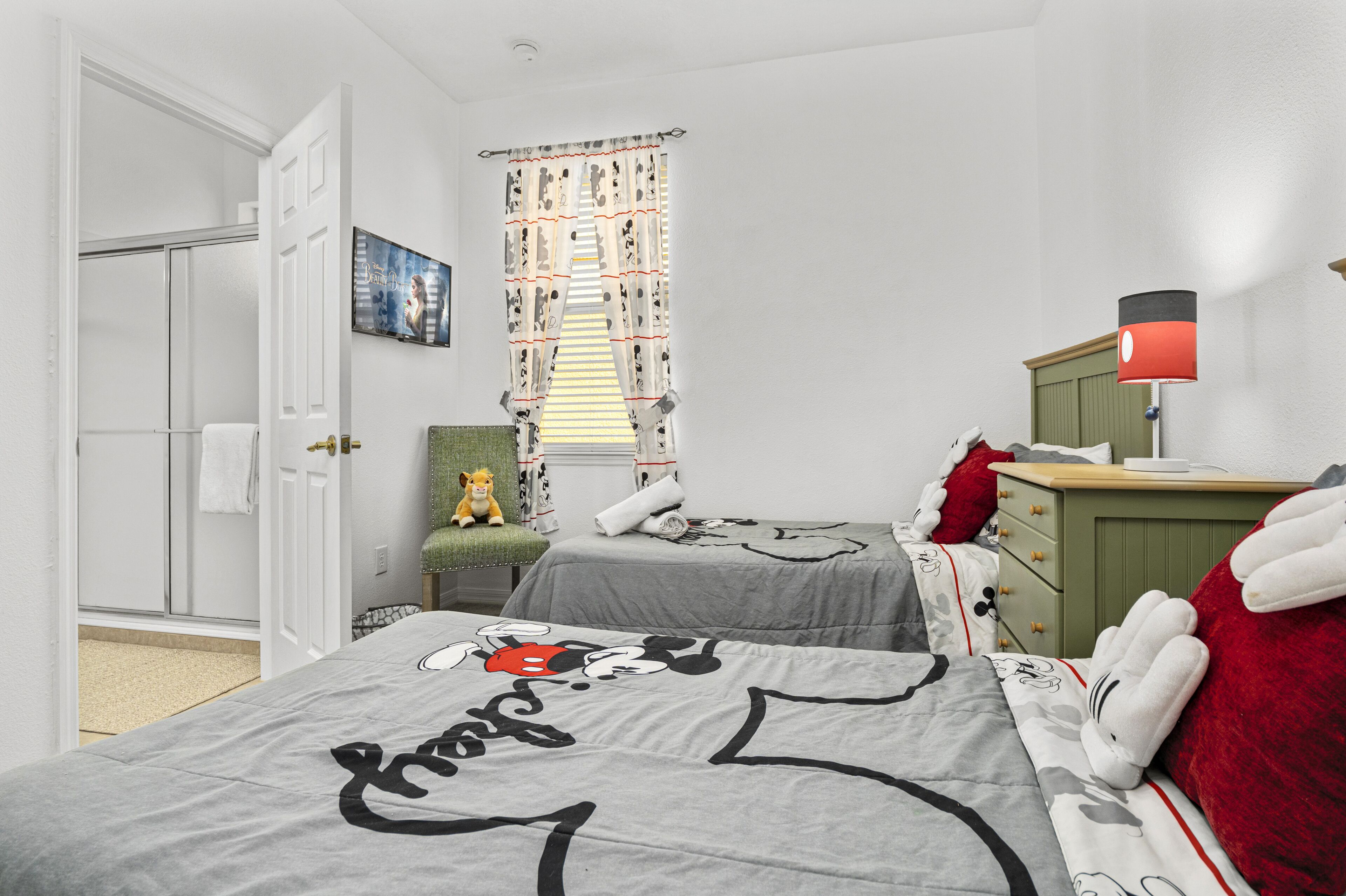 A bedroom with two beds, a green dresser, a lamp, a window with patterned curtains, a teddy bear, and a painting on the wall.