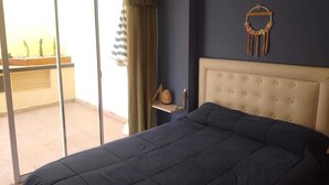 A bedroom with a large bed, a sliding glass door to a balcony, a small shelf with decorative items, and a wall hanging. - Cómodo, Amplio y Seguro Apartamento Familiar a Pasos del Metro en Buenos Aires (Buenos Aires)