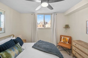 Un dormitorio con cama, una silla, cómoda, ventana con cortinas y ventilador de techo.
