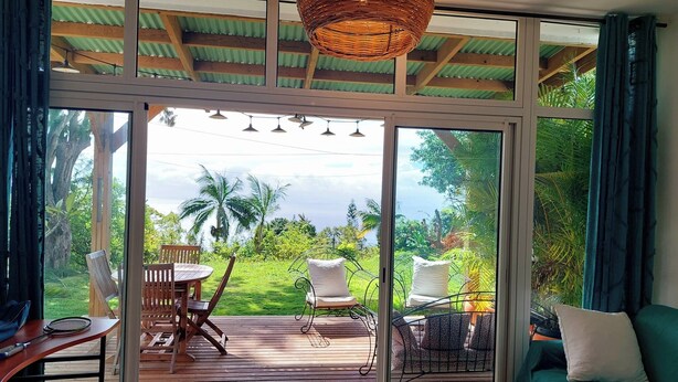 A room with a large sliding glass door leading to a deck with outdoor seating and a view of lush greenery. - Chez Lauret, à la Montagne (LA MONTAGNE)