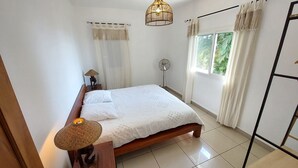 A neatly arranged bedroom with a bed, bedside lamp, and a standing fan. - Chez Lauret, à la Montagne (LA MONTAGNE)